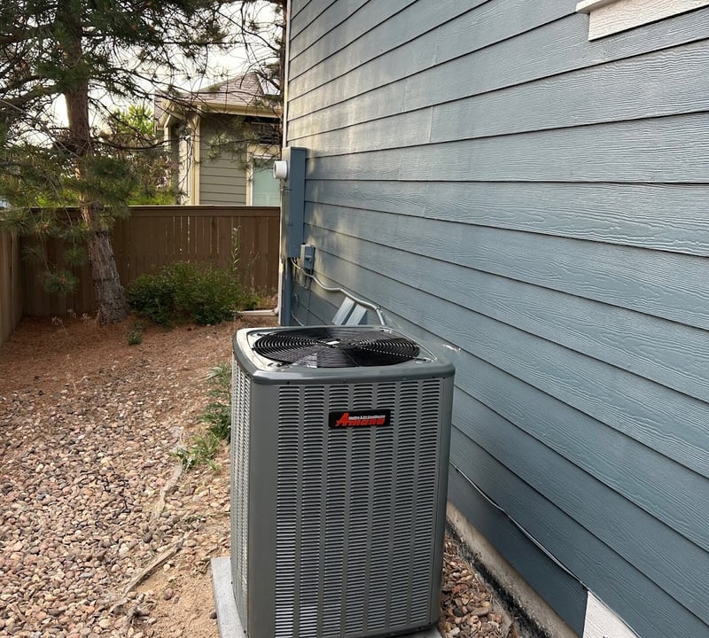Amana outdoor AC unit installed on a compressor pad beside a home with blue lap siding.