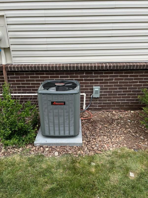 New Amana outdoor air-conditioning condenser installed on a concrete pad against the brick foundation of a home.