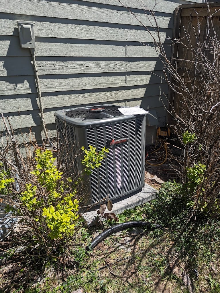 New Amana outdoor AC condenser installed on a pad in a fenced side yard between shrubs.