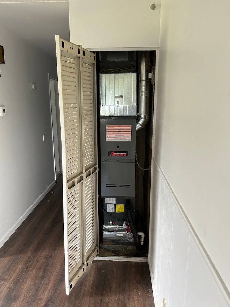 New Amana gas furnace installed in a hallway closet behind louvered bifold doors, above a hardwood floor.