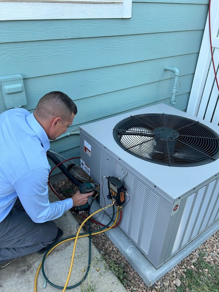 Colorado Bear technician kneeling next to a Rheem outdoor AC condenser, connecting refrigerant gauges to charge the system.