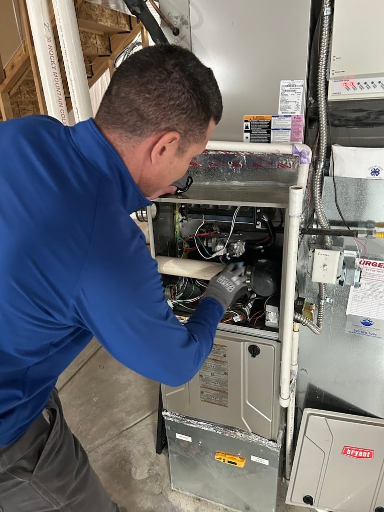 Colorado Bear technician in a blue uniform servicing an open gas furnace inside a home mechanical room.